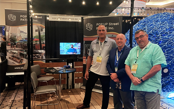 Three men with tags in front of their Pop Up Street Patio booth and display at a conference.