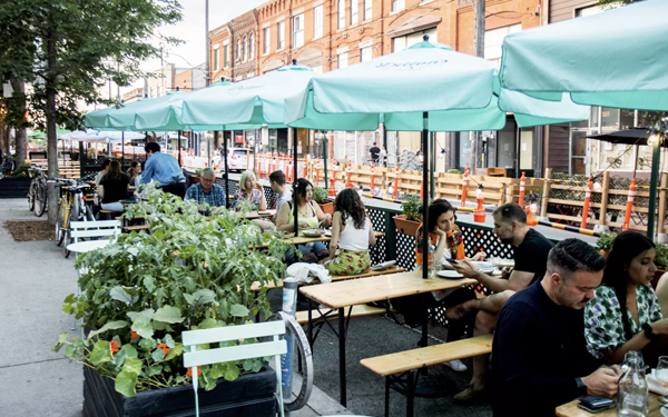 People sitting on the street patio, with light blue umbrellas and bright foliage planters.
